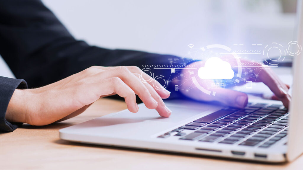 Manos escribiendo en una laptop con un ícono de nube y elementos digitales, simbolizando infraestructura en la nube.